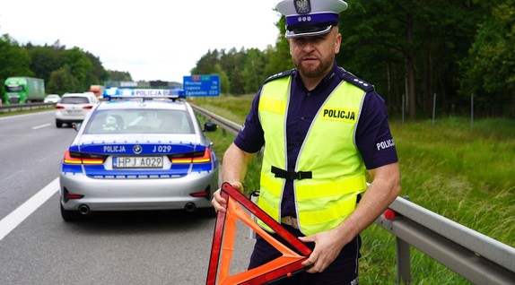 Policjant z trójkątem, przed radiowozem.