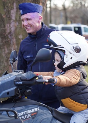 Policjant stoi obok motocykla, obok kobieta z dzieckiem.