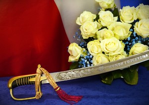Zdjęcie: szabla z napisem : Ojczyzna i honor na tle biało czerwonej flagi i białych róż.