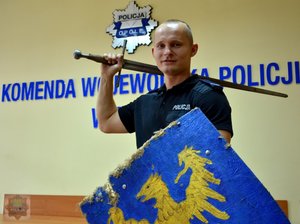 sierż. Kistela na tle napisu Komenda Wojewódzka Policji w Opolu z mieczem i tarczą w rękach