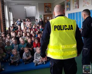Dzieci siedzą na podłodze i przysłuchują się wykładowi policjanta, część z nich ma podniesioną rękę do góry na znak zgłaszania się do odpowiedzi.