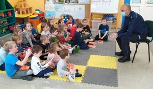 policjant w sali przedszkolnej rozdaje odblaski dzieciom siedzącym na dywanie
